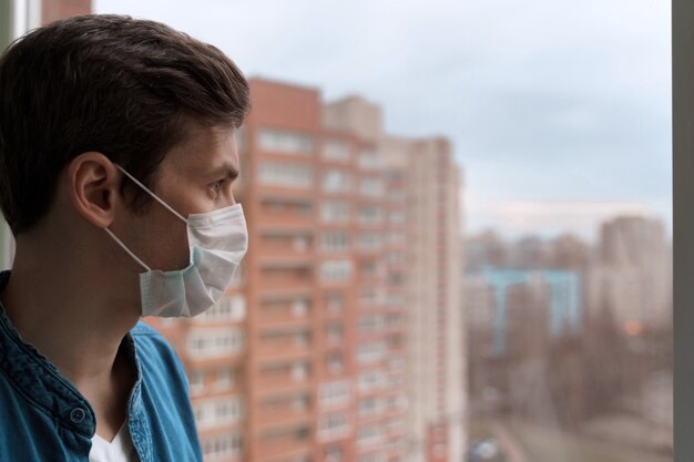 Jeune caucasien principal portant un masque de protection restant à l'isolement à la maison pour l'auto-quarantaine, regardant par la fenêtre.