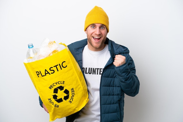 Jeune caucasien tenant un sac plein de bouteilles en plastique à recycler isolé sur fond blanc célébrant une victoire en position de vainqueur