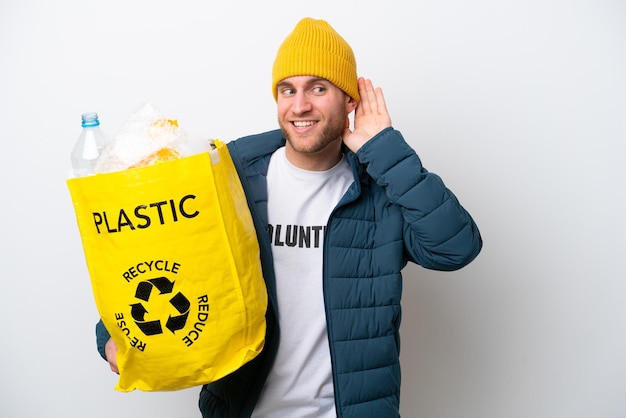 Jeune caucasien tenant un sac plein de bouteilles en plastique à recycler isolé sur fond blanc écoutant quelque chose en mettant la main sur l'oreille