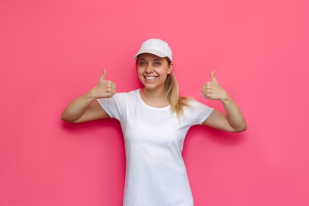 Une jeune caucasienne belle femme blonde souriante heureuse dans un t-shirt blanc et une casquette montrant le pouce vers le haut comme un geste avec deux mains isolées sur un mur rose de couleur vive