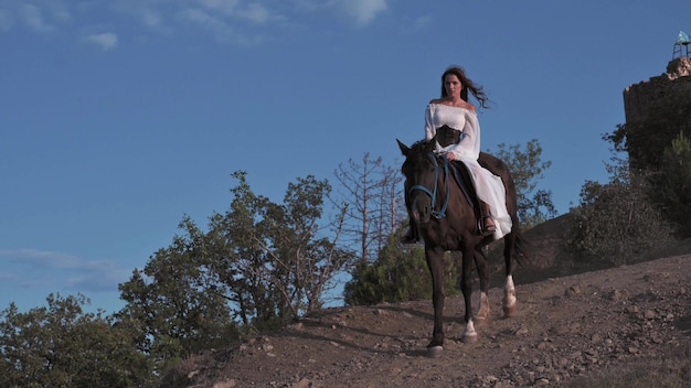 Une jeune cavalière vêtue d'une robe blanche descend de la pente à cheval sur fond d'un magnifique paysage de montagne aventure liberté 4k vidéo au ralenti