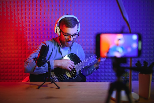 Un jeune chanteur caucasien dans un casque tient un nouveau single de guitare sur un smartphone au home studio Un artiste du millénaire utilise un instrument de musique pour chanter un clip vidéo sur un téléphone portable