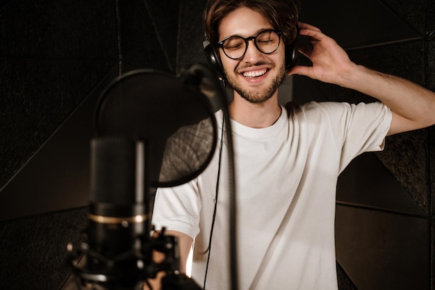 Jeune chanteur masculin attrayant dans des écouteurs chantant joyeusement dans un studio de son moderne