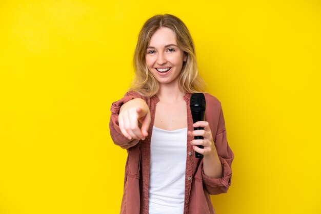 Jeune chanteuse caucasienne prenant un microphone isolé sur fond jaune surpris et pointant vers l'avant