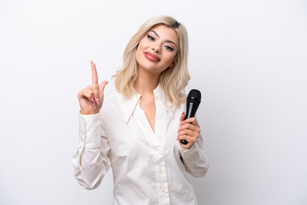 Jeune chanteuse femme prenant un microphone isolé sur fond blanc montrant et levant un doigt en signe du meilleur