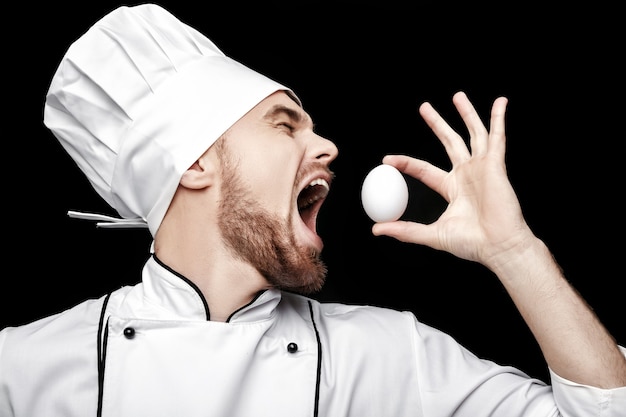 Jeune chef barbu en uniforme blanc détient un oeuf sur fond noir