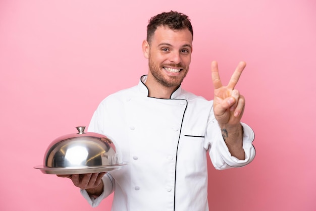 Jeune chef brésilien avec plateau isolé sur fond rose souriant et montrant le signe de la victoire