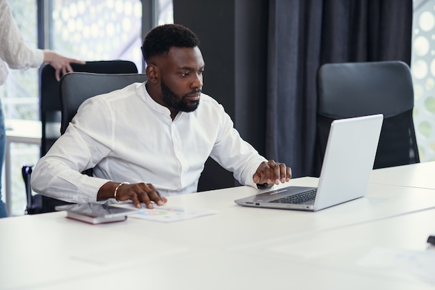 Un jeune chef de bureau à la peau foncée travaille avec différents rapports et un ordinateur portable de table sur son lieu de travail dans la salle de réunion du centre d'affaires moderne.