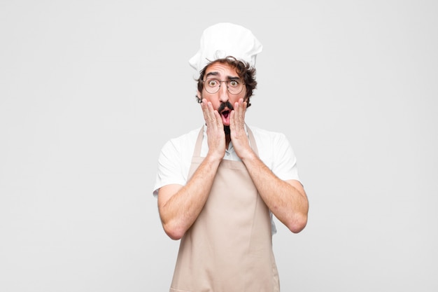 Jeune chef fou se sentir choqué et effrayé, terrifié avec la bouche ouverte et les mains sur les joues contre le mur blanc