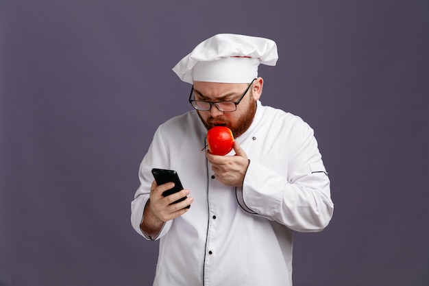 Jeune chef fronçant les sourcils portant des lunettes uniformes et une casquette à l'aide de son téléphone portable tout en mangeant une pomme isolée sur fond violet