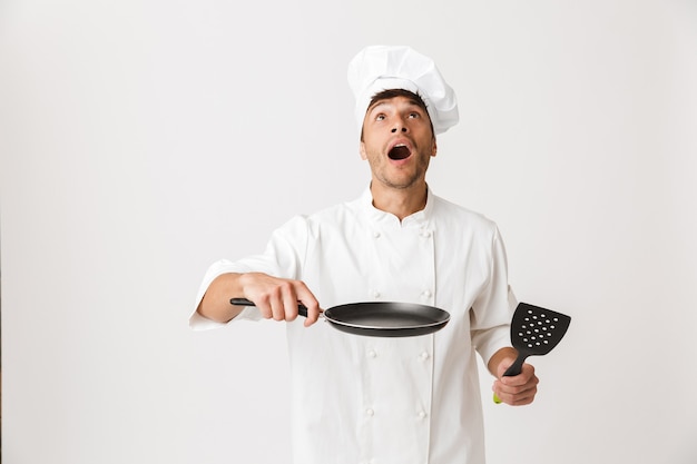 Jeune chef homme debout isolé sur un mur blanc tenant une poêle à frire.