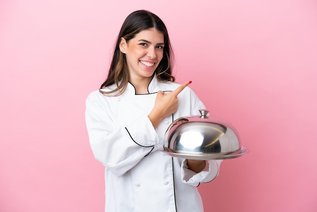 Jeune chef italien femme tenant un plateau avec couvercle isolé sur fond rose pointant le doigt sur le côté