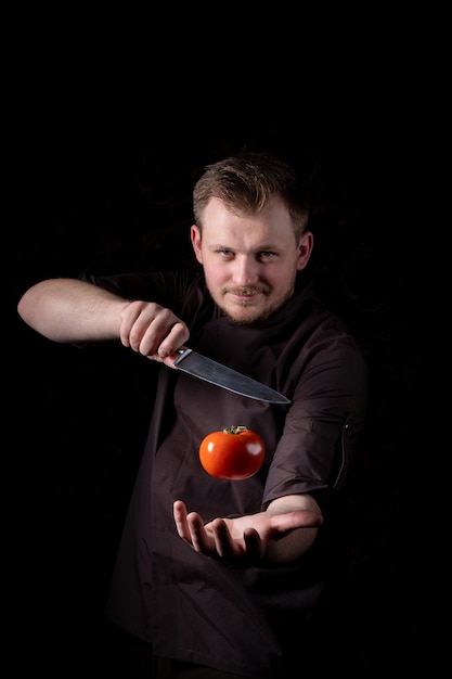 Un jeune chef masculin avec un couteau de chef professionnel attrape une tomate rouge fraîche sur fond noir