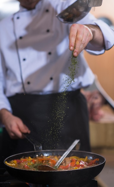 Jeune chef masculin mettant des épices sur des légumes dans un wok dans une cuisine commerciale