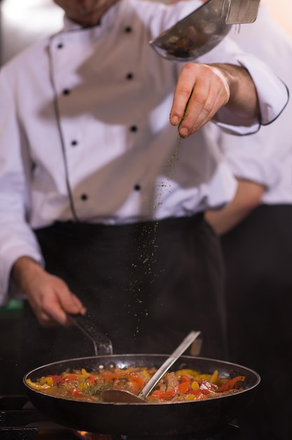 Jeune chef masculin mettant des épices sur des légumes dans un wok dans une cuisine commerciale