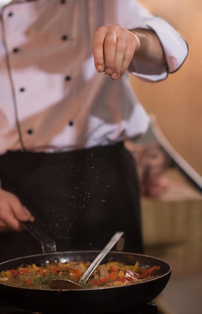 Jeune chef masculin mettant des épices sur des légumes dans un wok dans une cuisine commerciale