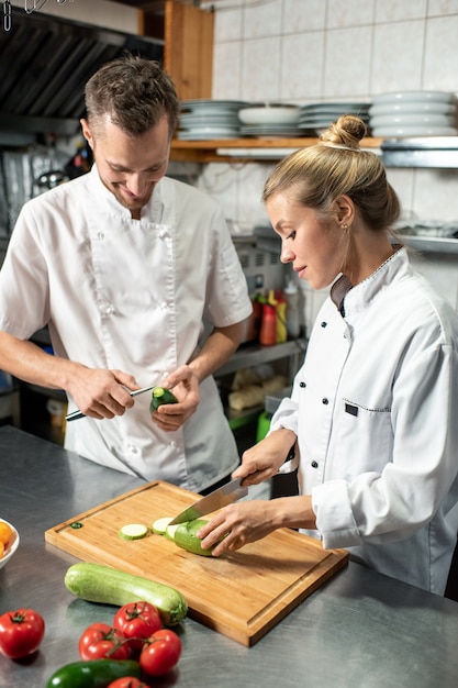 Jeune chef masculin professionnel avec un couteau épluchant l'avocat tandis que sa stagiaire coupe des courgettes fraîches sur une planche de bois dans la cuisine