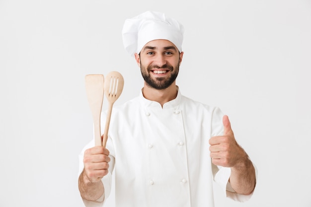 jeune chef en uniforme de cuisinier souriant tout en tenant des ustensiles de cuisine en bois isolés sur un mur blanc