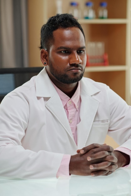 Jeune chercheur ou clinicien sérieux en blouse de laboratoire se concentrant sur ce qu'il regarde en s'asseyant