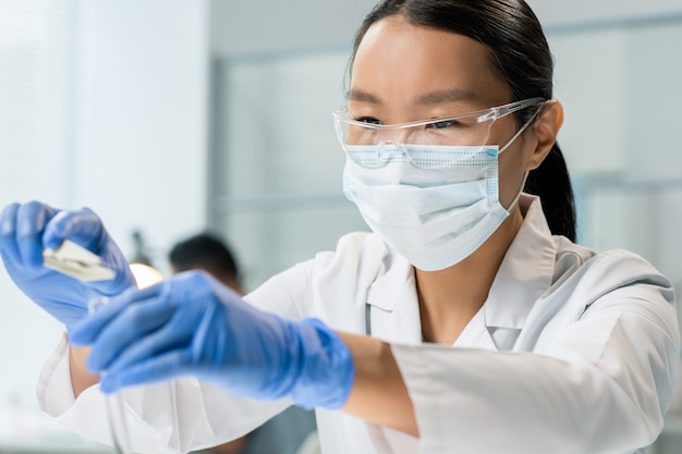 Jeune chercheuse gantée en blouse blanche et masque mettant un petit échantillon d'ingrédient alimentaire dans un flacon pendant une expérience en laboratoire