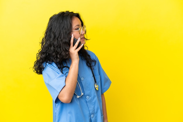 Jeune chirurgien médecin femme asiatique isolée sur fond jaune gardant une conversation avec le téléphone portable avec quelqu'un