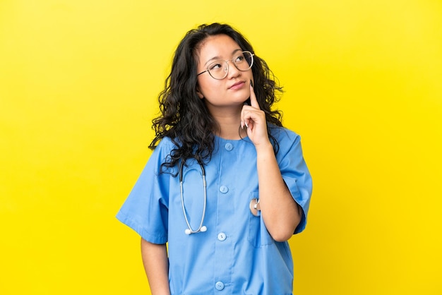 Jeune chirurgien médecin femme asiatique isolée sur fond jaune pensant à une idée tout en levant les yeux