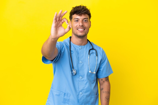 Jeune chirurgien médecin homme isolé sur fond jaune montrant signe ok avec les doigts