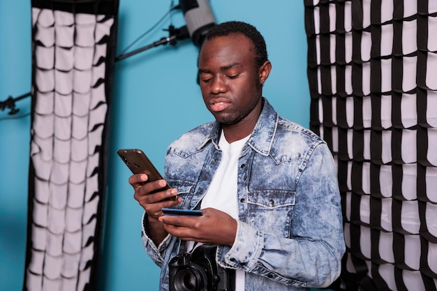 Jeune client utilisant une carte de débit pour le paiement en ligne tout en se tenant dans un studio de production avec un équipement photo professionnel. Homme avec appareil photo reflex numérique payant sans espèces sur un magasin de détail numérique.