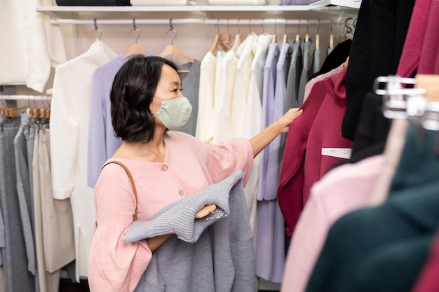 Jeune cliente asiatique en masque et chemisier élégant rose debout près d'un rack avec un nouvel assortiment de vêtements décontractés et choisissant un pull