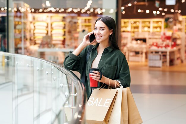 Jeune cliente réussie avec boisson et sacs en papier parlant sur téléphone mobile après avoir visité les départements du grand centre commercial moderne