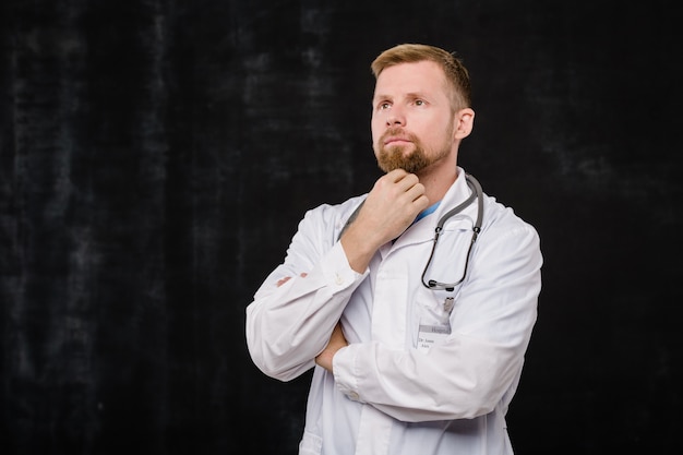 Jeune clinicien barbu avec expression pensive debout dans l'isolement sur fond noir