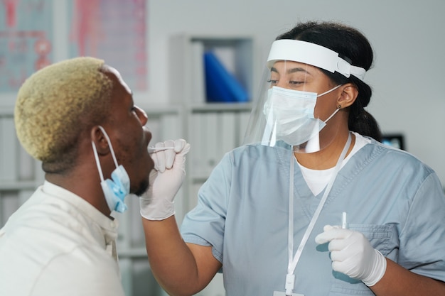 Jeune clinicienne métisse debout devant un patient africain avec la bouche ouverte tout en le testant pour covid dans les cliniques