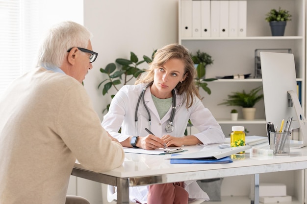 Une jeune clinicienne sérieuse en blouse blanche qui regarde un patient âgé malade.