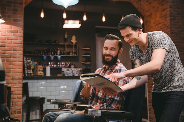 Jeune Coiffeur Heureux Et Beau Client De Contenu Regardant Le Magazine Assis Dans Un Salon De Coiffure