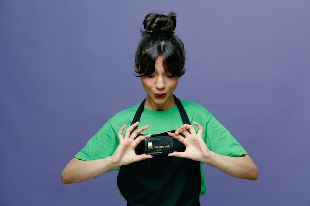 Jeune coiffeuse femme portant un tablier tenant une carte de crédit à la recherche d'un sourire heureux et positif souriant joyeusement debout sur fond bleu