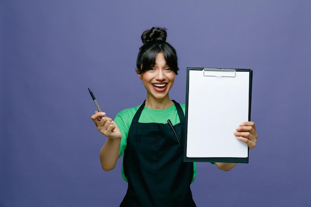 Jeune coiffeuse femme portant un tablier tenant un presse-papiers et un stylo regardant la caméra heureuse et joyeuse souriant largement debout sur fond bleu