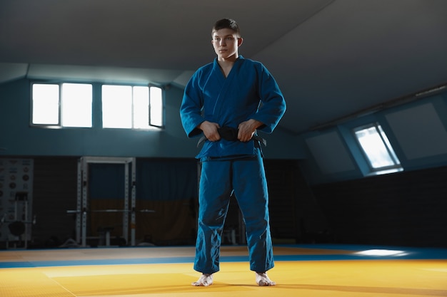 Jeune combattant caucasien de judo en kimono bleu avec ceinture noire posant confiant dans la salle de sport, fort et en bonne santé.