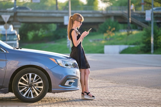 Jeune conductrice debout près de sa voiture parlant sur un téléphone portable dans une rue de la ville en été
