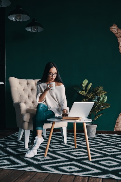 Jeune et confiant. Belle jeune femme utilisant un ordinateur portable et tenant une tasse assise dans le fauteuil