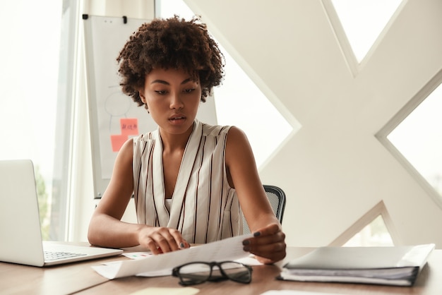 Photo jeune connaisseur. belle femme afro-américaine analysant des documents alors qu'elle était assise dans un bureau moderne. concept d'entreprise. la vie de bureau