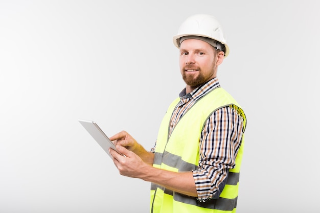 Jeune constructeur barbu à succès en casque blanc, chemise à carreaux et gilet jaune à l'aide de tablette devant la caméra