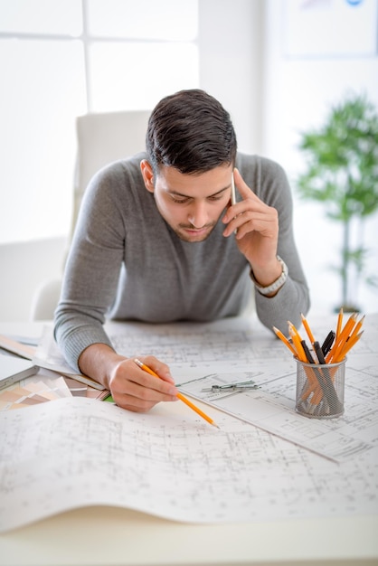 Jeune constructeur masculin arabe analysant le plan au bureau au bureau et utilisant le téléphone.