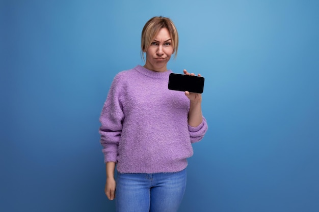 Jeune consultante blonde en détresse en sweat à capuche violet tenant un smartphone avec maquette sur bleu