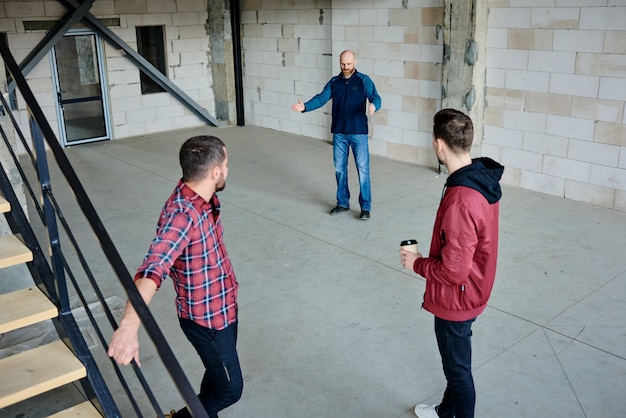 Photo jeune contremaître confiant en tenue décontractée expliquant à ses collègues où il est juste de faire un mur divisant une grande pièce en deux plus petites