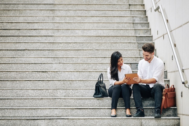 Jeune couple d'affaires multiethnique souriant assis sur des marches et discutant d'un document sur une tablette