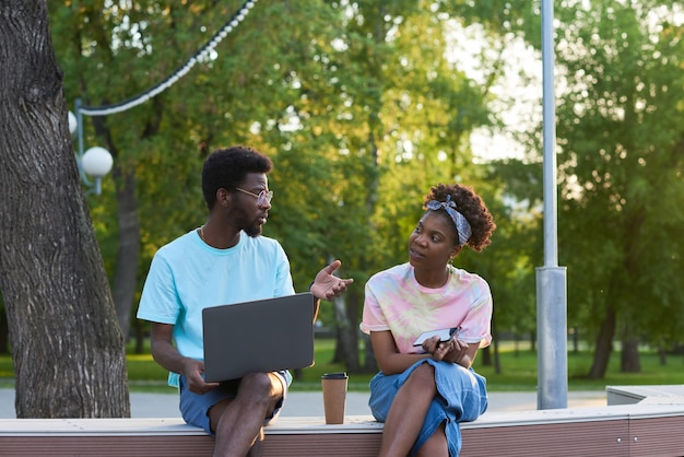 Jeune couple africain travaillant sur un ordinateur portable et discutant du travail en ligne ensemble tout en étant assis à l'extérieur