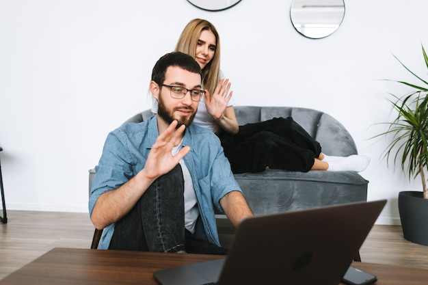 Jeune couple à l'aide d'un ordinateur portable en regardant l'écran du portable sur appel vidéo et salutation avec agitant dans le salon moderne.