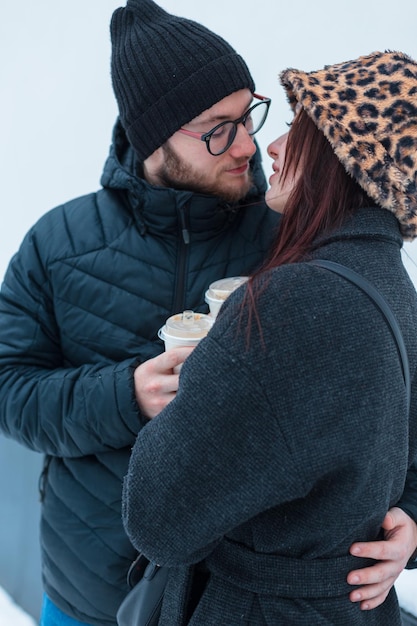 Un jeune couple d'amants à la mode en vêtements d'hiver avec une veste tiennent une tasse de café et s'embrassent sur un fond blanc