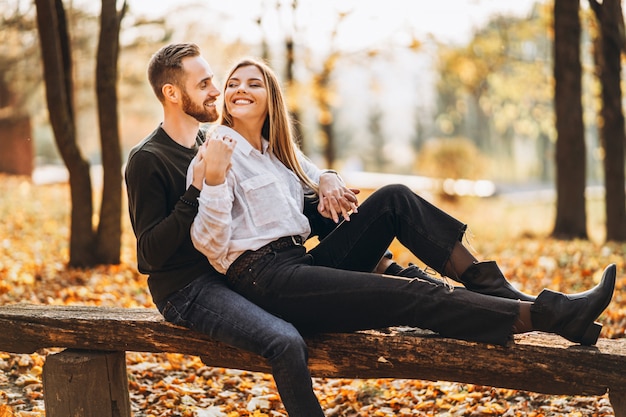 Un jeune couple d'amoureux assis sur un banc en bois dans la forêt. Homme femme, étreindre, et, sourire, sur, les, fond, de, arbres automne