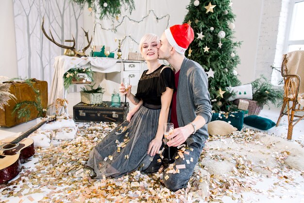 Jeune couple amoureux assis sur le sol près de l'arbre de Noël jetant des confettis dorés